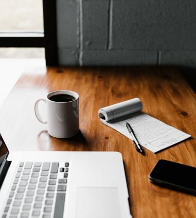 Drewniany stół, na nim laptop, kawa, notes z długopisem i smartfon